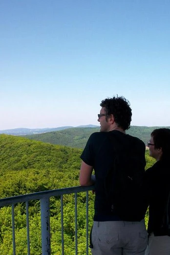 Zwei Personen betrachten von einer Aussichtsplattform aus die weiten, bewaldeten Hügel einer Landschaft.