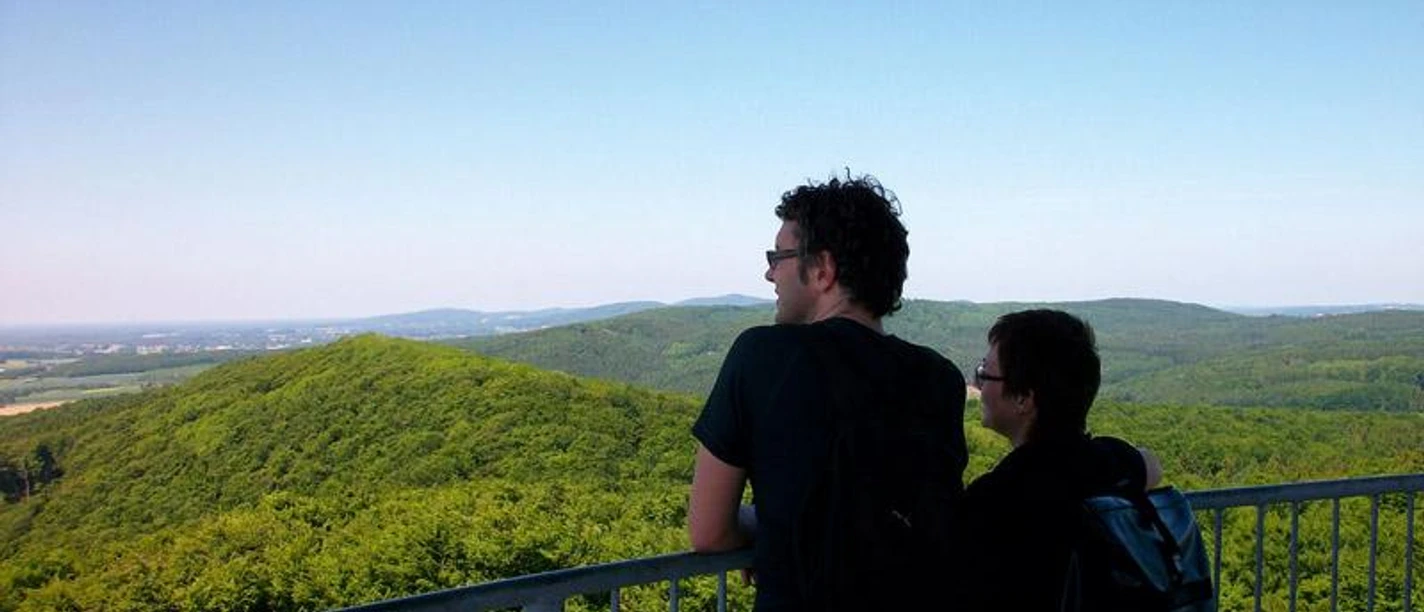Die Aussicht genießen Zwei Personen betrachten von einer Aussichtsplattform aus die weiten, bewaldeten Hügel einer Landschaft.