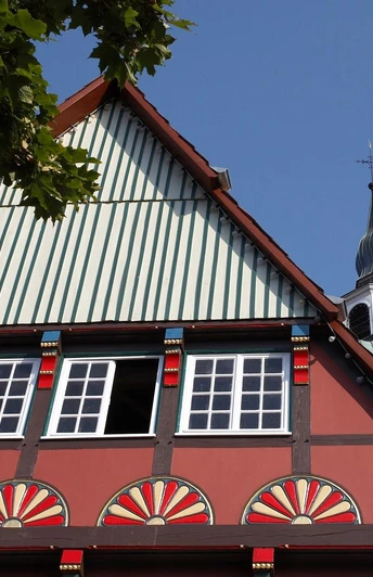 Fachwerkhaus mit bunten Ornamenten, im Hintergrund Kirchturm unter blauem Himmel sichtbar.