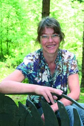 Marijke Smit Eine Frau mittleren Alters mit Brille sitzt lächelnd in einem grünen Wald und trägt ein gemustertes Hemd.