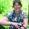 Marijke Smit Eine Frau mittleren Alters mit Brille sitzt lächelnd in einem grünen Wald und trägt ein gemustertes Hemd.