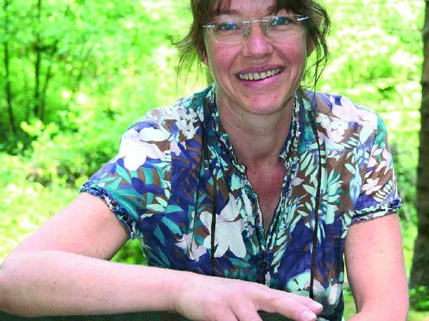 Marijke Smit Eine Frau mittleren Alters mit Brille sitzt lächelnd in einem grünen Wald und trägt ein gemustertes Hemd.