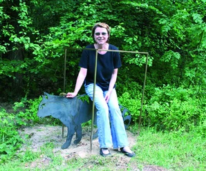 Birgit Schwarz-Kalkreuter Eine Frau sitzt lächelnd auf einer Skulptur einer Katze aus Metall in einem grünen Park.