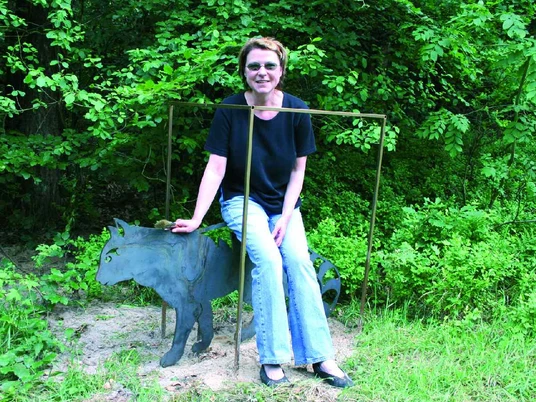 Birgit Schwarz-Kalkreuter Eine Frau sitzt lächelnd auf einer Skulptur einer Katze aus Metall in einem grünen Park.