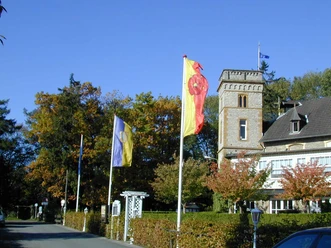 Historisches Gebäude mit Turm, umgeben von herbstlichen Bäumen, beflaggt mit bunten Fahnen.