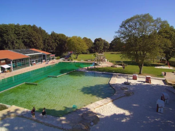 Freibad mit großem Schwimmbecken, umliegenden Liegewiesen und einem Spielplatz im Hintergrund.
