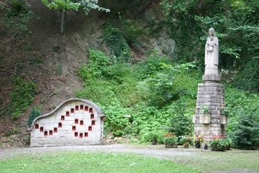 Marienskulptur Eine Marienskulptur steht neben einer Steinwand mit kleinen, rot eingetäfelten Nischen im Grünen.
