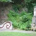 Marienskulptur Eine Marienskulptur steht neben einer Steinwand mit kleinen, rot eingetäfelten Nischen im Grünen.
