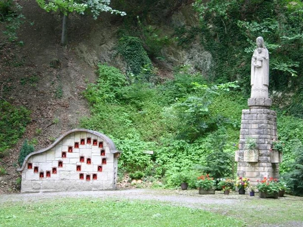 Marienskulptur Eine Marienskulptur steht neben einer Steinwand mit kleinen, rot eingetäfelten Nischen im Grünen.