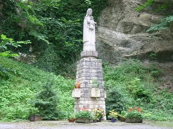 Marienskulptur Steinskulptur der Maria mit Jesuskind auf hohem Sockel, umgeben von blühenden Pflanzen im Grünen.