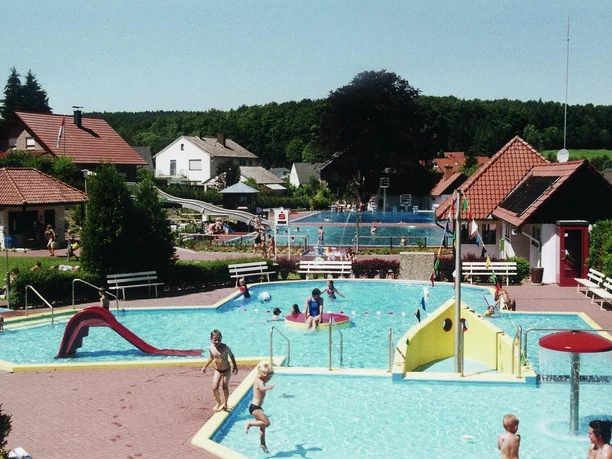 Waldschwimmbad Preußisch Oldendorf Kinderbecken mit Rutsche und Wasserspielen vor einem Waldschwimmbad an einem sonnigen Tag.