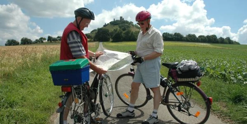 Zwei Radfahrer studieren eine Karte vor einem bewachsenen Hügel mit einer Ruine an der Spitze.