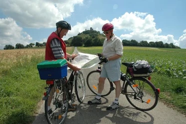 Der Desenberg: Wahrzeichen des Warburger Landes Zwei Radfahrer studieren eine Karte vor einem bewachsenen Hügel mit einer Ruine an der Spitze.