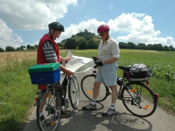 Der Desenberg: Wahrzeichen des Warburger Landes Zwei Radfahrer studieren eine Karte vor einem bewachsenen Hügel mit einer Ruine an der Spitze.