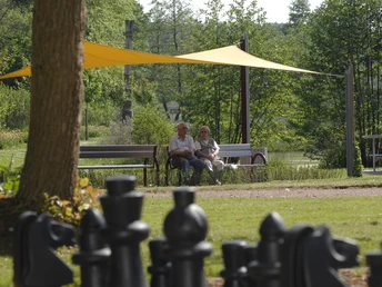 Zwei ältere Personen sitzen auf einer Bank im Kurpark, umgeben von Bäumen und einem großen Schachbrett.