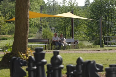 Zwei ältere Personen sitzen auf einer Bank im Kurpark, umgeben von Bäumen und einem großen Schachbrett.