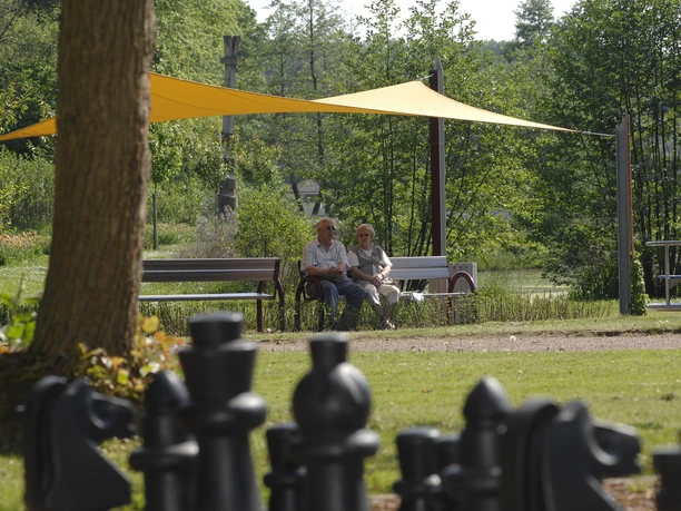 Schatten- und Charakterplatz im Kurpark Zwei ältere Personen sitzen auf einer Bank im Kurpark, umgeben von Bäumen und einem großen Schachbrett.