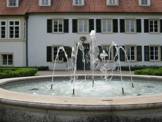 Springbrunnen vor dem Haus des Gastes Ein mehrstrahliger, runder Springbrunnen vor einem zweistöckigen Haus mit schwarzen Fensterläden.