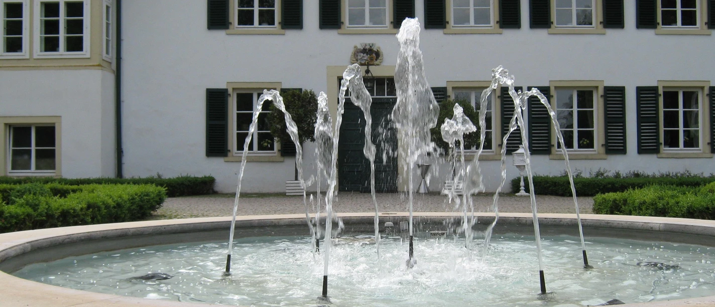 Springbrunnen vor dem Haus des Gastes Ein mehrstrahliger, runder Springbrunnen vor einem zweistöckigen Haus mit schwarzen Fensterläden.