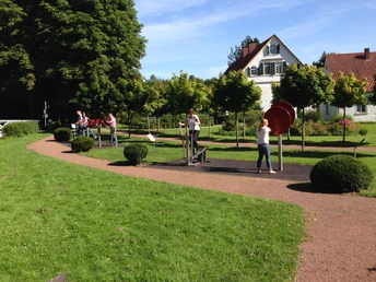Menschen nutzen Fitnessgeräte im Garten mit grüner Wiese, Bäumen und einem weißen Gebäude im Hintergrund.