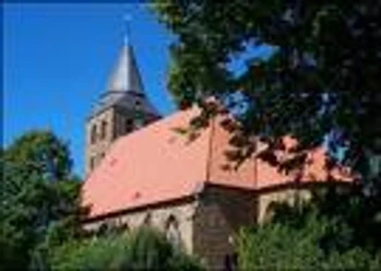 Ev. Kirche Bad Holzhausen Backsteinkirche mit rotem Dach und spitzem Turm, umgeben von Bäumen unter strahlend blauem Himmel.