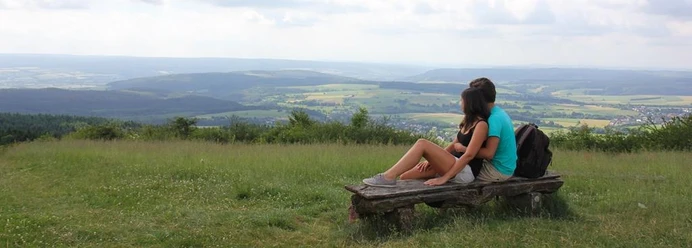Ausblick vom Köterberg Zwei Personen sitzen auf einer Bank und blicken über eine weitläufige, grüne Hügellandschaft.