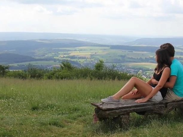 Ausblick vom Köterberg Zwei Personen sitzen auf einer Bank und blicken über eine weitläufige, grüne Hügellandschaft.