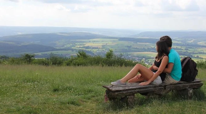 Ausblick vom Köterberg Zwei Personen sitzen auf einer Bank und blicken über eine weitläufige, grüne Hügellandschaft.