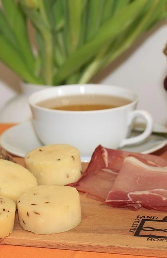 Brot, Käse mit Kümmel und Schinken auf Holzbrett, begleitet von Kaffeetasse und Trauben.
