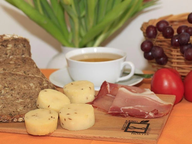Brot, Käse und Schinken - Leckereien aus Nieheim Brot, Käse mit Kümmel und Schinken auf Holzbrett, begleitet von Kaffeetasse und Trauben.