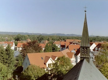 Kath. Kirche St. Ursula 1 Kirchturmspitze von St. Ursula inmitten eines Wohngebiets mit roten Dächern und umgebender Natur.