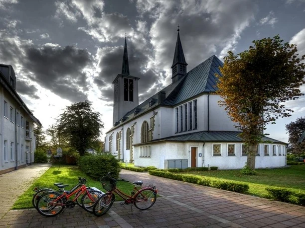 Katholische Kirche St. Ursula mit markanten Türmen, umgeben von Bäumen und parkenden Fahrrädern.