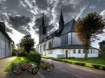 Kath. Kirche St. Ursula 3 Katholische Kirche St. Ursula mit markanten Türmen, umgeben von Bäumen und parkenden Fahrrädern.