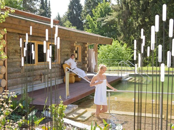 Zwei Personen in Handtüchern entspannen am holzverkleideten Naturbadeteich mit angrenzender Terrasse.