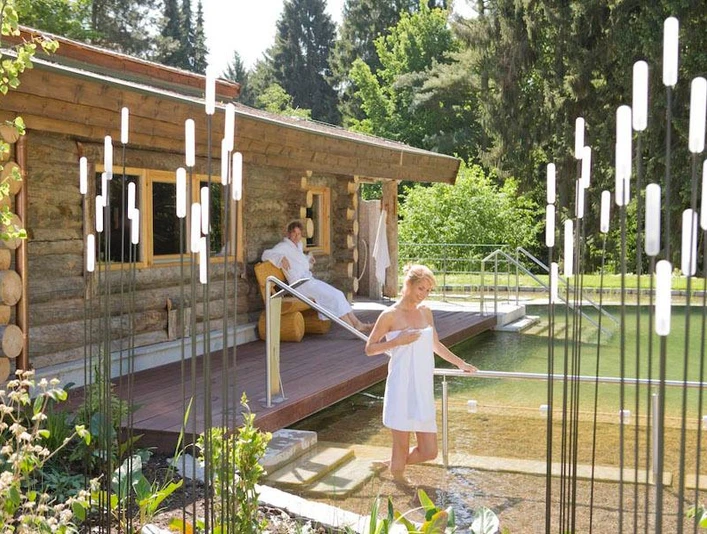 Zwei Personen in Handtüchern entspannen am holzverkleideten Naturbadeteich mit angrenzender Terrasse.
