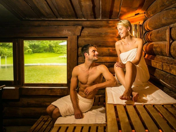 Eine Frau und ein Mann in Handtüchern entspannen in einer holzgetäfelten Sauna mit Blick auf einen Teich.