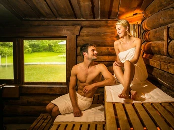 VitaSol Therme: SeeSauna im SaunaPark mit angrenzendem Naturbadeteich Eine Frau und ein Mann in Handtüchern entspannen in einer holzgetäfelten Sauna mit Blick auf einen Teich.