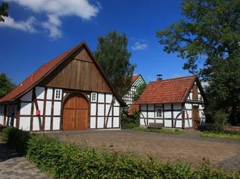 Bockhorster Kotten & Backhaus Fachwerkhäuser unter blauem Himmel mit Ziegeln gedeckt, umgeben von grünen Bäumen und Hecken.