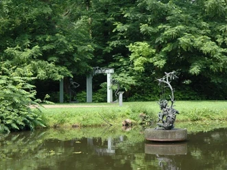 Bronzeskulptur auf Teich mit grünen Bäumen und einer Gartenlaube im Hintergrund.
