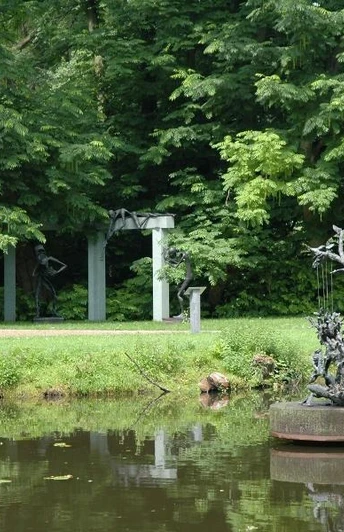 Bronzeskulptur auf Teich mit grünen Bäumen und einer Gartenlaube im Hintergrund.
