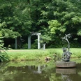 Klostergarten Rietberg - Skulpturenpark Wilfried Koch Bronzeskulptur auf Teich mit grünen Bäumen und einer Gartenlaube im Hintergrund.