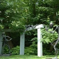 Skulpturen im Garten zeigen stilisierte menschliche Figuren aus Metall vor üppigem grünem Hintergrund.