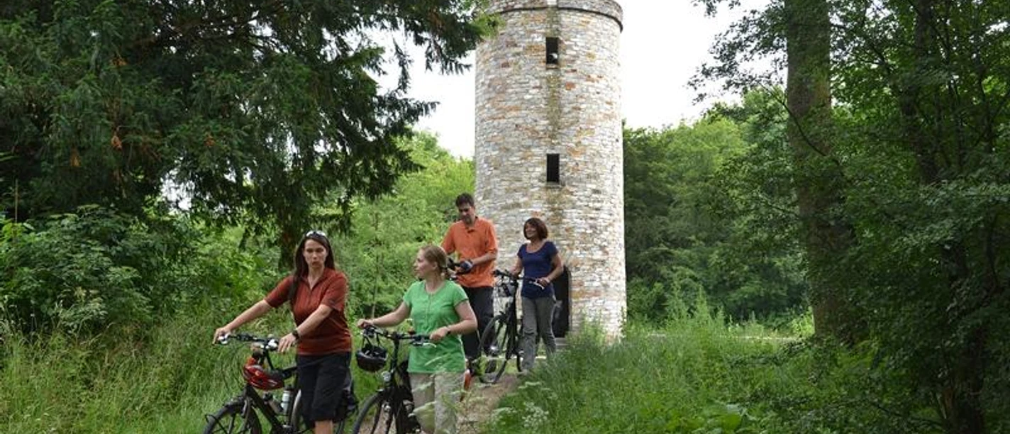 Radfahrergruppe passiert einen historischen Turm, umgeben von üppigem Grün im Sommer.