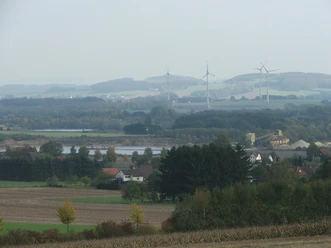 Blick auf Kiesteiche mit umliegenden Feldern, Windrädern und sanften Hügeln im Hintergrund.