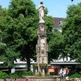 Eine steinerne Mariensäule auf einem Platz, umgeben von Laubbäumen, mit Passanten im Vordergrund.