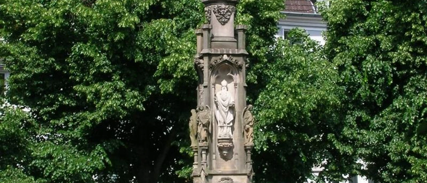 Eine steinerne Mariensäule auf einem Platz, umgeben von Laubbäumen, mit Passanten im Vordergrund.