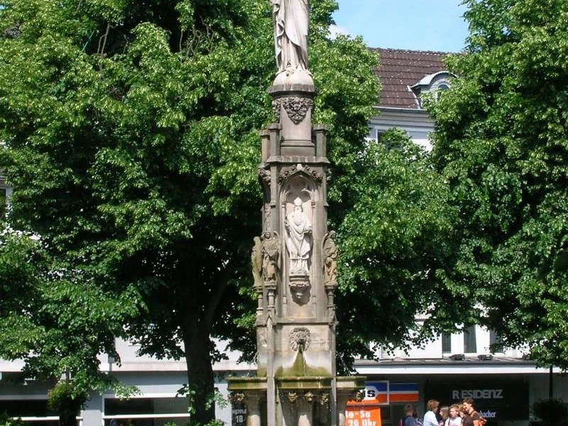 Eine steinerne Mariensäule auf einem Platz, umgeben von Laubbäumen, mit Passanten im Vordergrund.