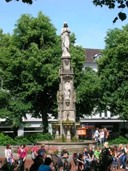 Mariensäule auf dem Marienplatz Eine steinerne Mariensäule auf einem Platz, umgeben von Laubbäumen, mit Passanten im Vordergrund.