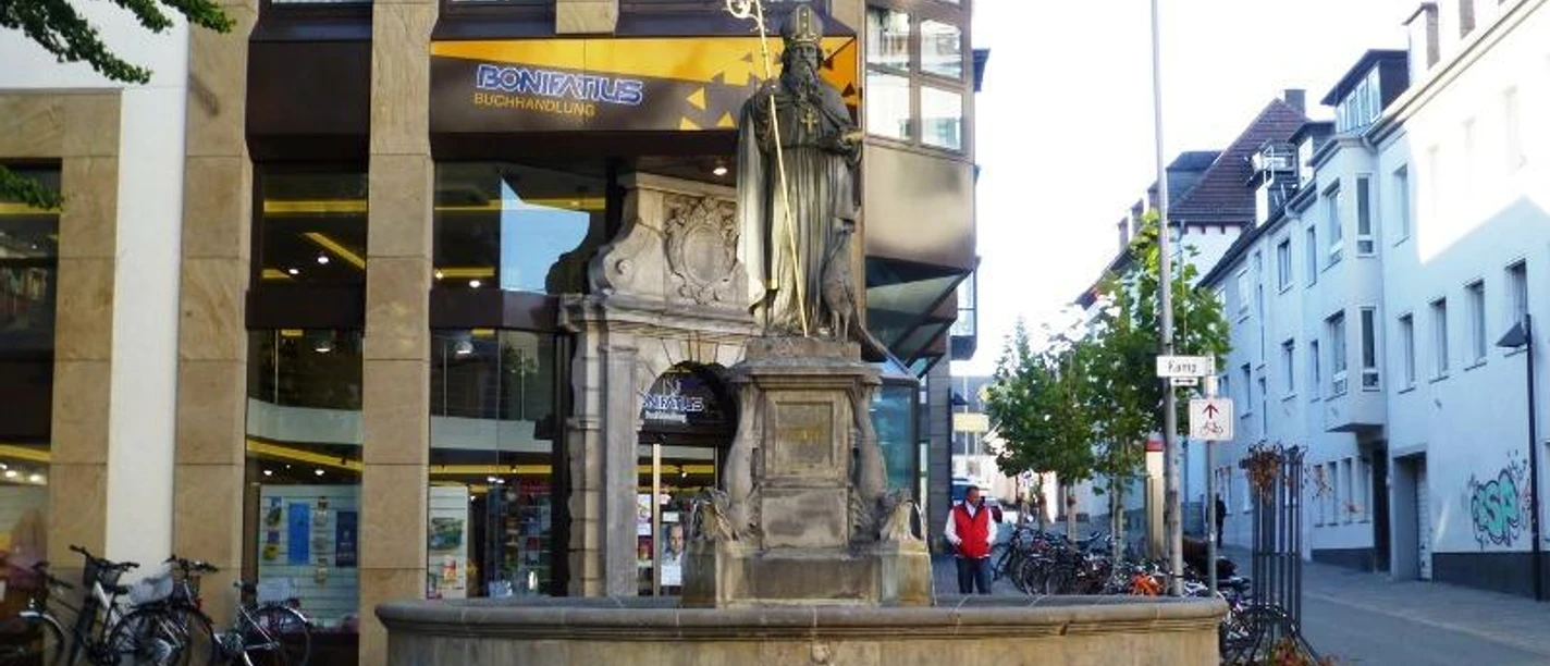 Brunnen mit Statue eines Heiligen vor einem modernen Gebäude; Fahrräder parken um den Brunnen.