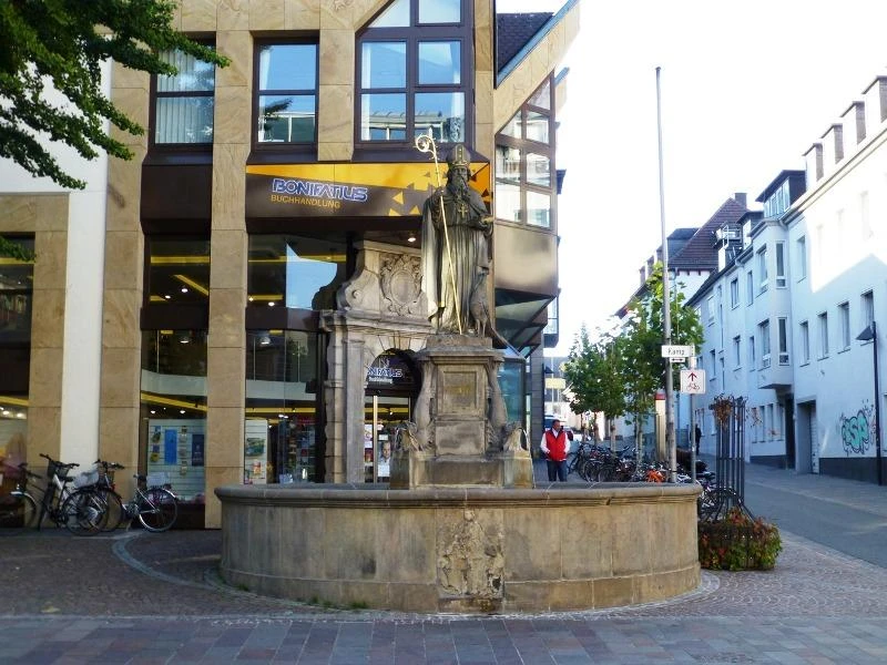 Brunnen mit Statue eines Heiligen vor einem modernen Gebäude; Fahrräder parken um den Brunnen.